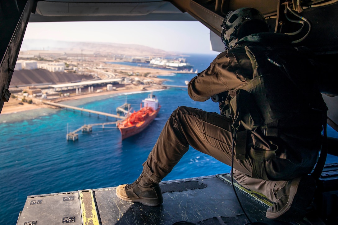 A Marine looks out the door of a helicopter at a ship in the water.