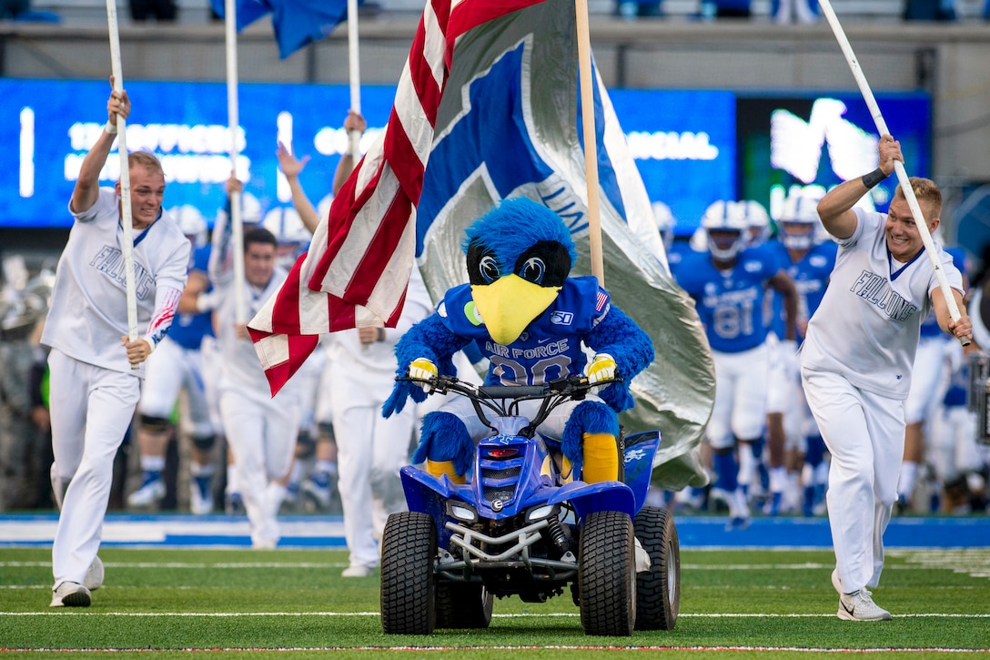 A bird mascot drives an all-terrain vehicle onto a football field, followed by athletes.