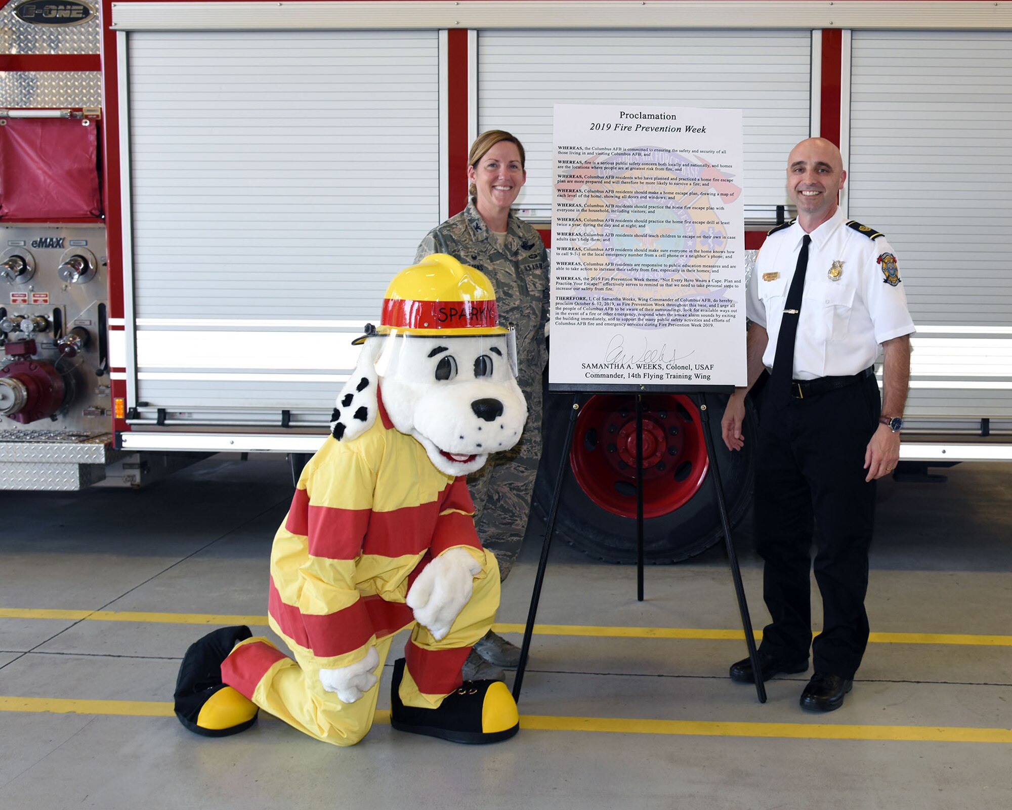 Col. Samantha Weeks, 14th Flying Training Wing commander, stands with Shawn Ricchuito, 14th Civil Engineering Squadron fire chief, and Sparky with the 2019 Fire Prevention Week proclamation, Sept 16, 2019, on Columbus Air Force Base, Miss. Columbus AFB will be hosting an open house and a parade on Oct. 5 to kick off Fire Prevention Week. (Courtesy Photo)