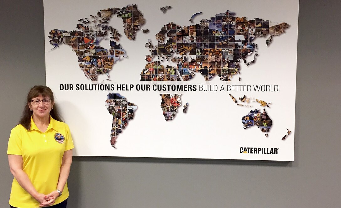 Woman in yellow DLA shirt stands in front of Caterpillar's map with the slogan, "Our solutions help our customers build a better world."