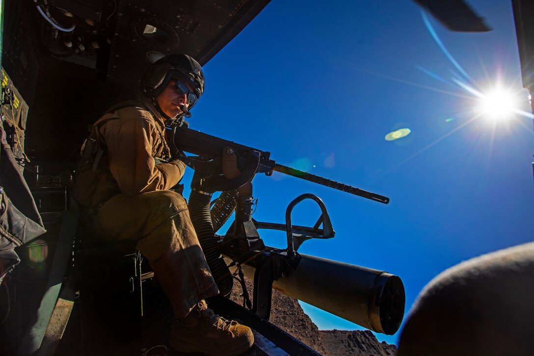 A Marine manning a machine gun aboard an open aircraft looks out onto vibrant blue sky.