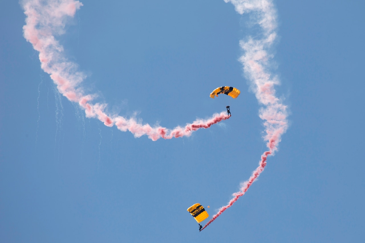 Two parachutists leave pink smoke in their wake as they perform against a blue sky.