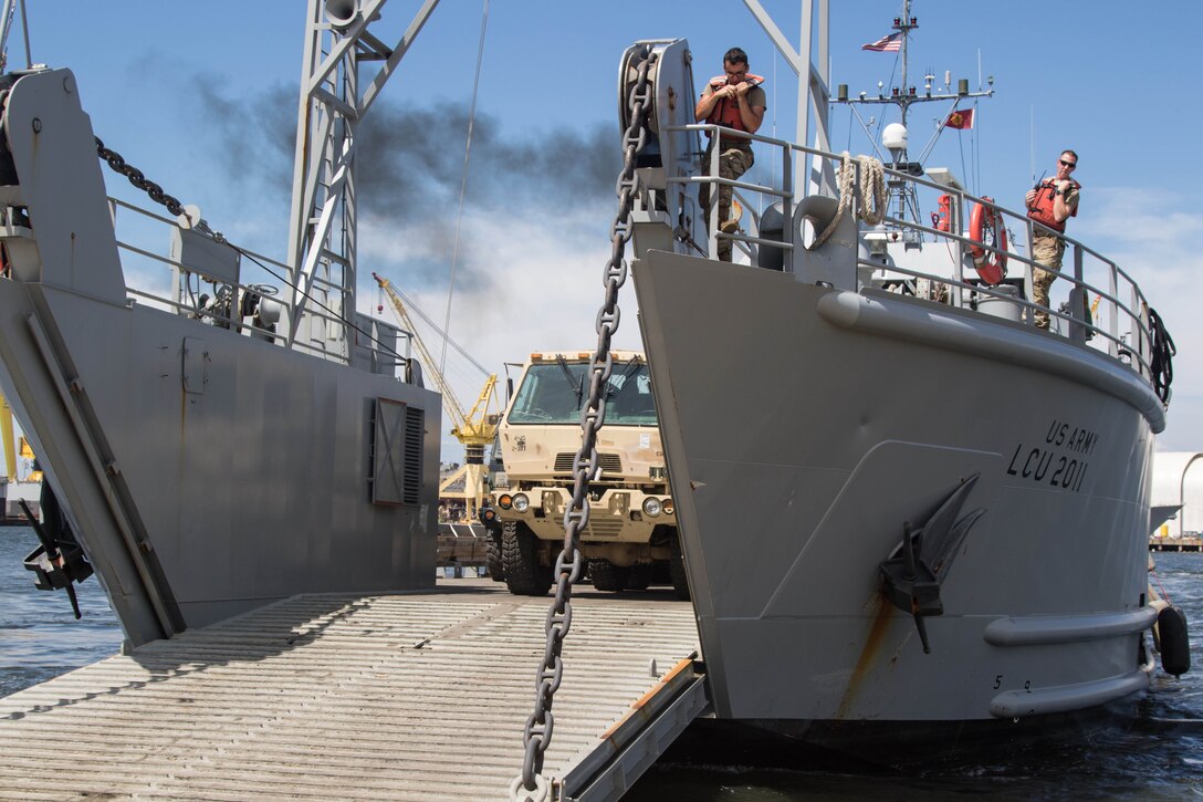 Soldiers aboard a boat and wearing floatation vests observe as a large tactical truck goes down the boat’s ramp.