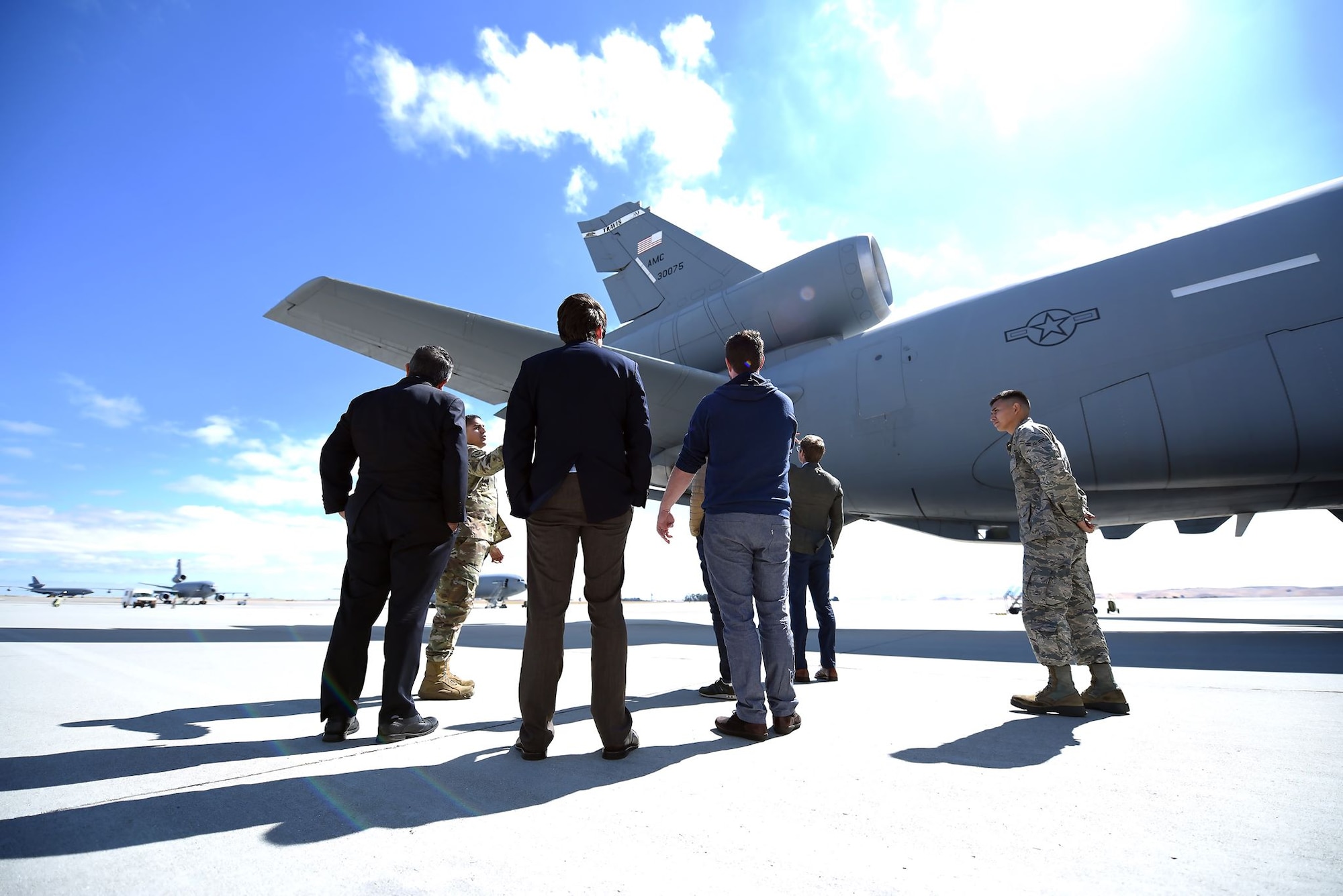 Men in suits along with men in military uniforms look out on the tail of a KC-10 Extender on a long expanse of runway
