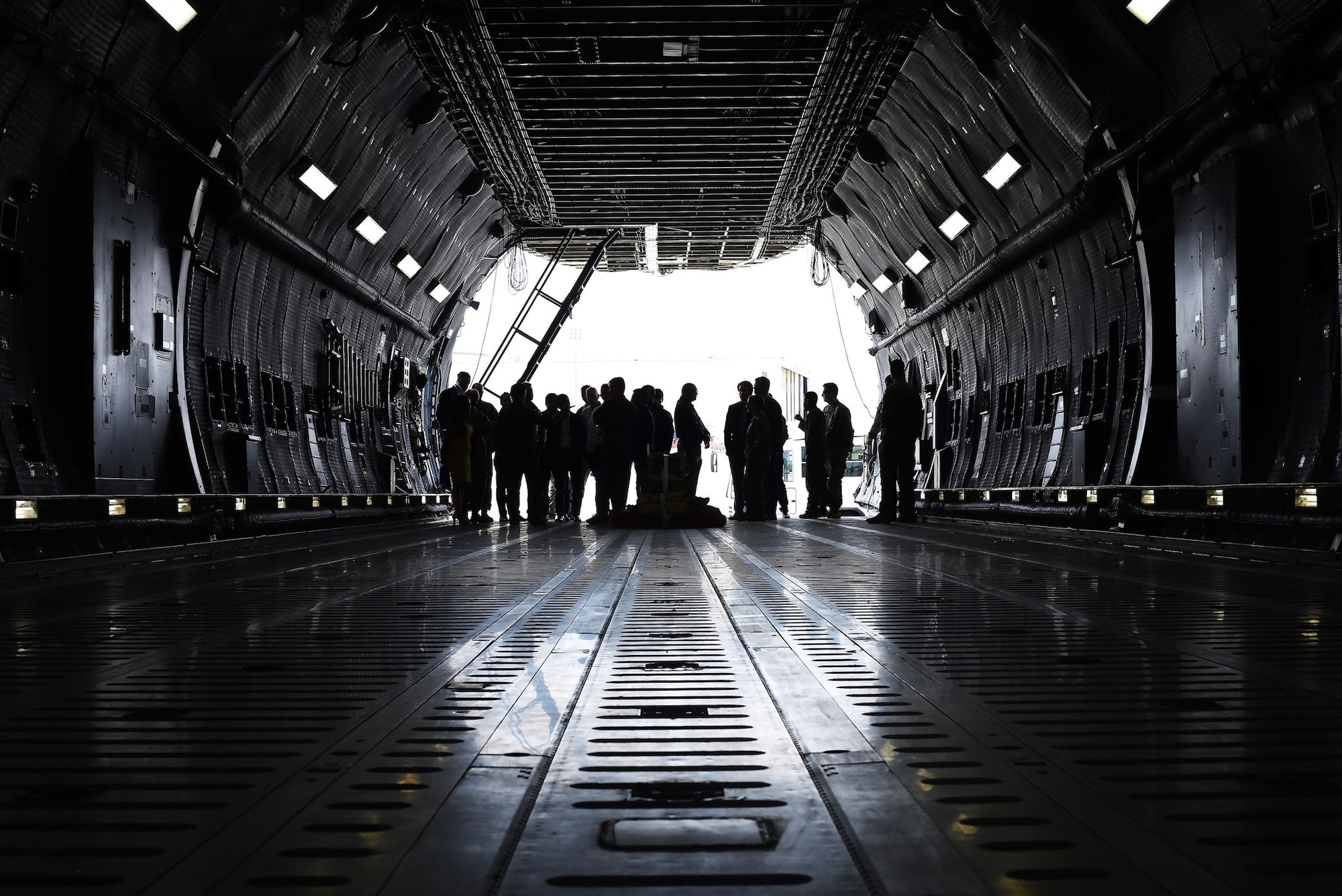 A gaggle of civilians and military members are silhouetted against the bright sunlight outside of the C-5M Super Galaxy in which they currently stand