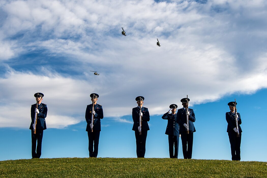 Six Air Force cadets stand in formation in a field as three aircraft fly over them.