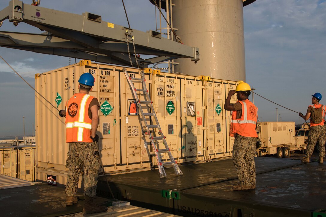 Sailors in protective helmets and orange vests hold guide lines attached to a crane that is preparing to lift a shipping container.