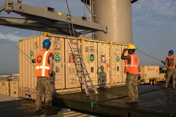 Sailors in protective helmets and orange vests hold guide lines attached to a crane that is preparing to lift a shipping container.