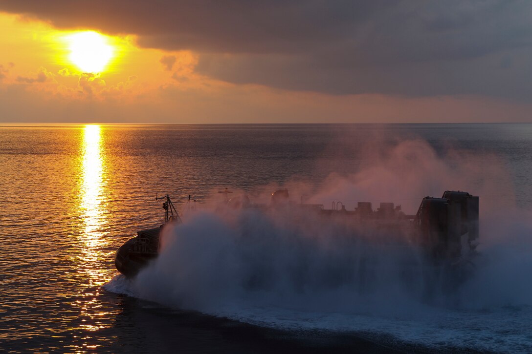 A military landing craft makes a big splash as it leaves a ship at twilight.