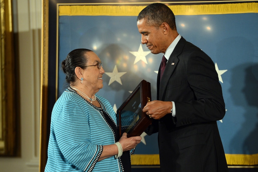 A woman wearing a dress and glasses smiles as she accepts the Medal of Honor, encased in a wooden box with a glass top, from former President Obama.