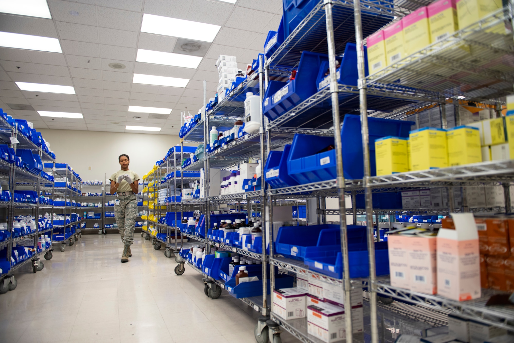 A pharmacy technician walks down an isle contain bottles medication on each side.