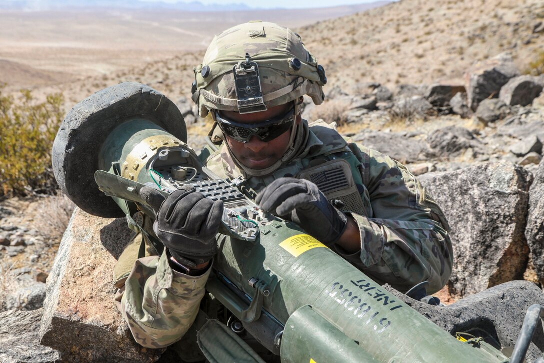 A soldier wearing gloves, a helmet and sunglasses handles a missile while out in desert-type terrain.