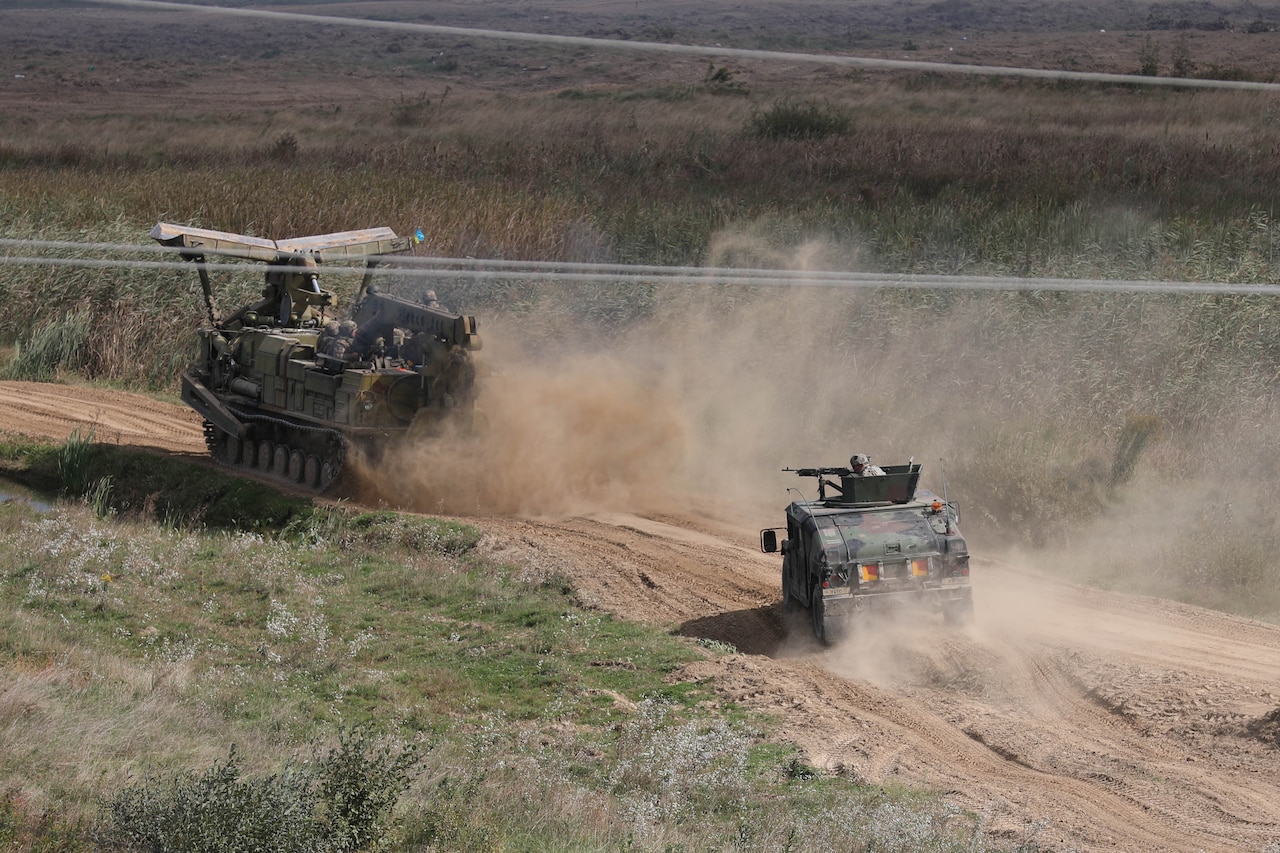 Two military combat vehicles traverse a dirt path.