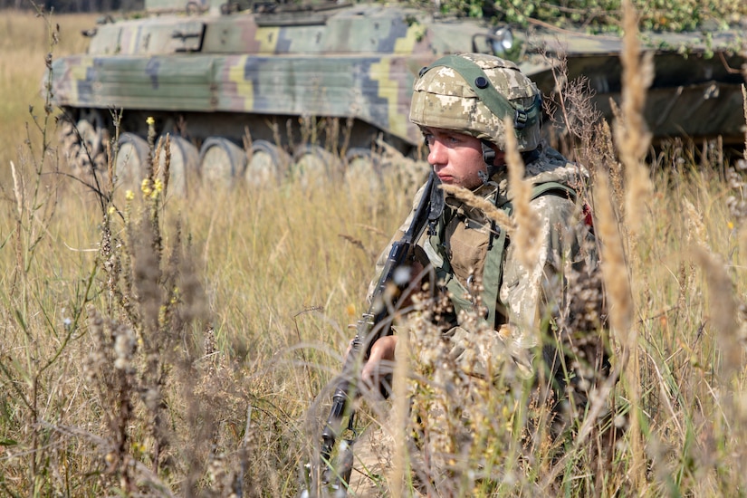 A soldier stands in a grassy area with his rifle pointed toward the ground. In the background is a large military vehicle.