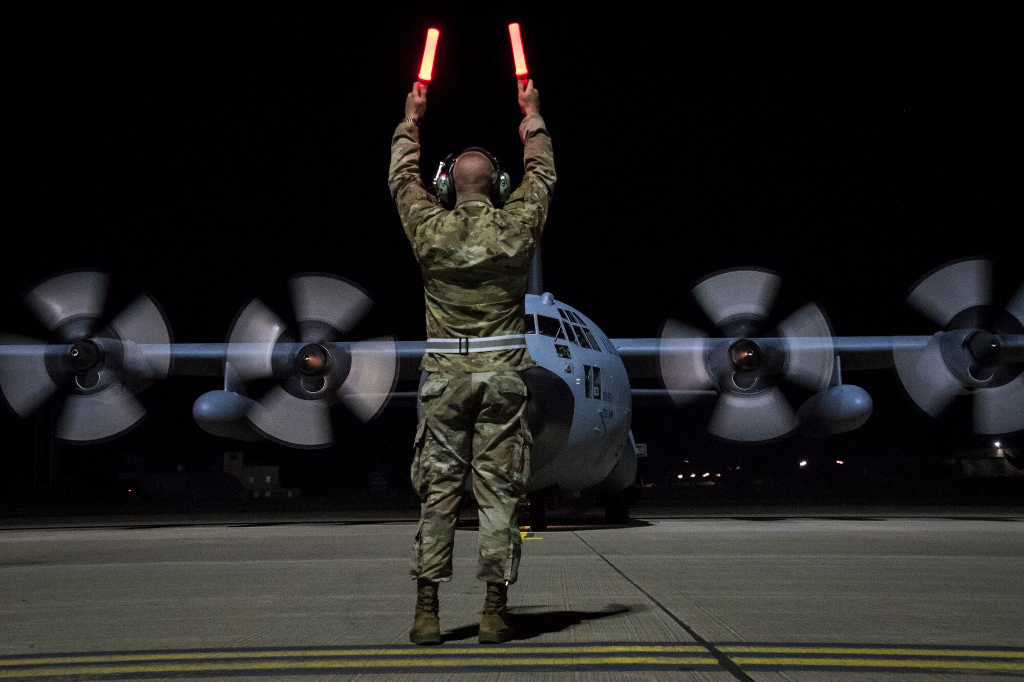 Photo of an Airman waving directions to a moving C-130 on the flight line during early morning.