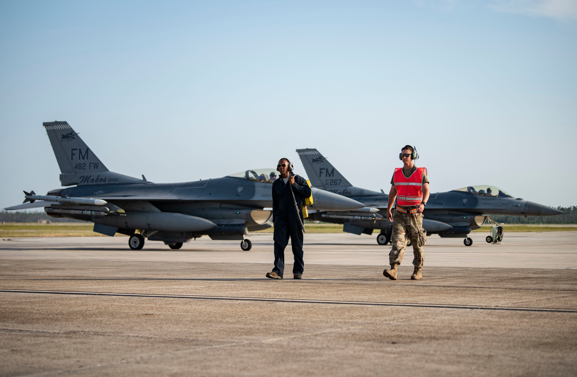 Two Airmen walk on the flight line with two aircraft behind them.