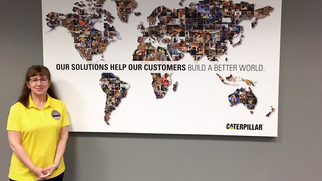 Woman in yellow DLA shirt stands in front of Caterpillar's map with the slogan, "Our solutions help our customers build a better world."