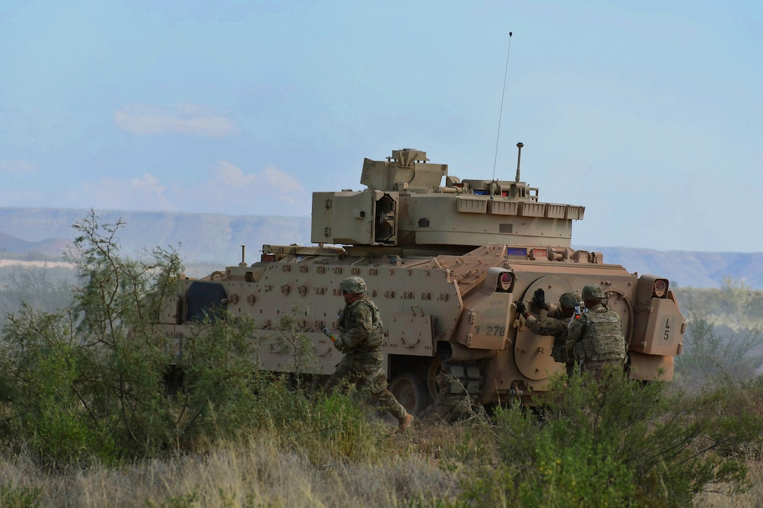 A tank rolls across an open field with several soldiers around it.