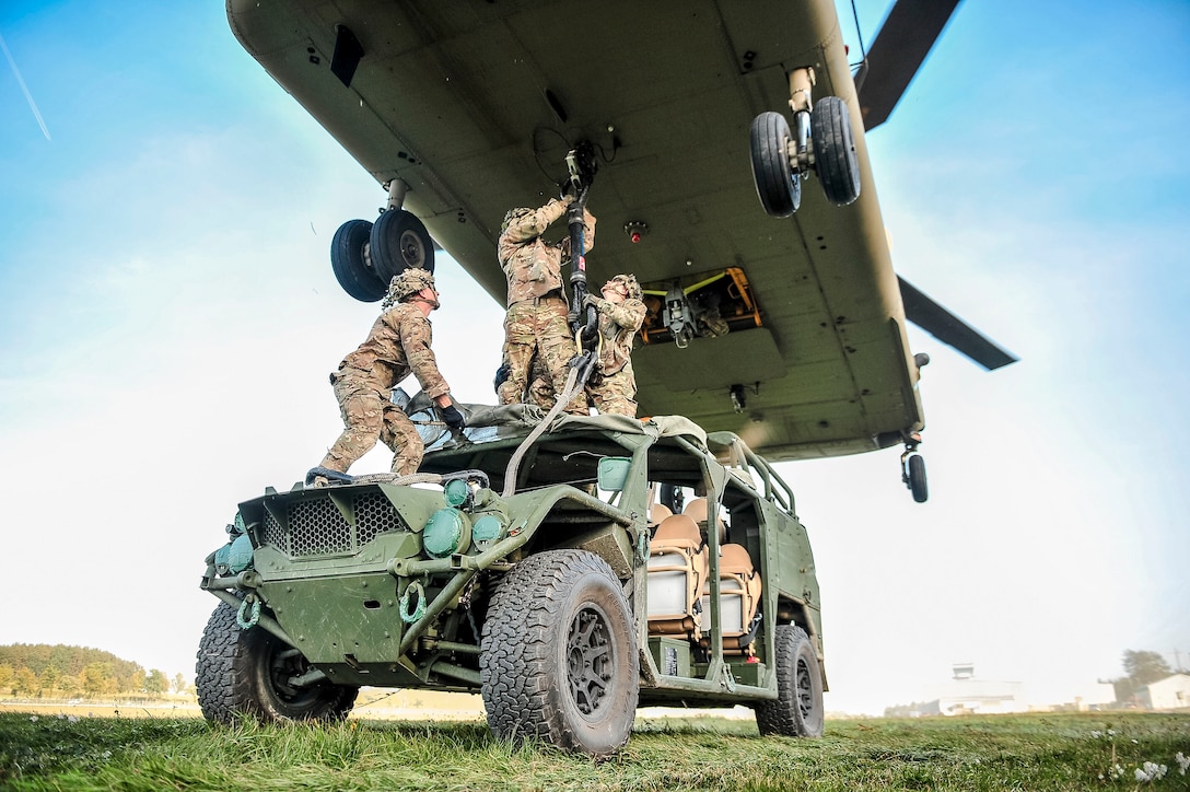 Soldiers stand on top of a tactical vehicle and attach it by sling to a helicopter's underbelly.