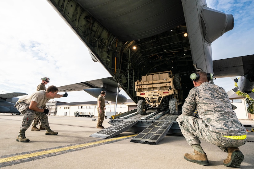 Four men in camouflage uniforms watch as an all-terrain vehicle backs out of  an aircraft.