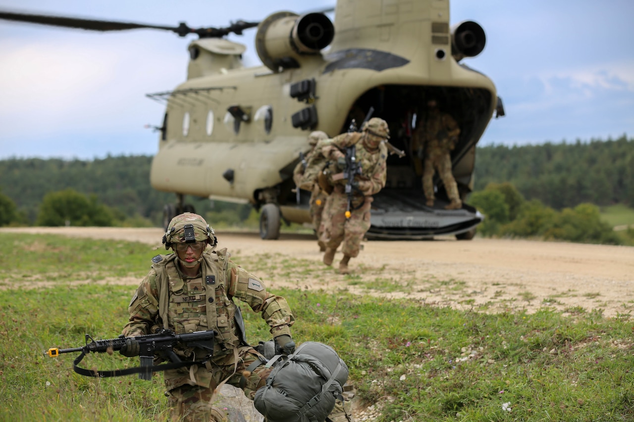 Troops in camouflage uniforms scramble out of helicopter as one soldier crouches on the ground with a weapon.