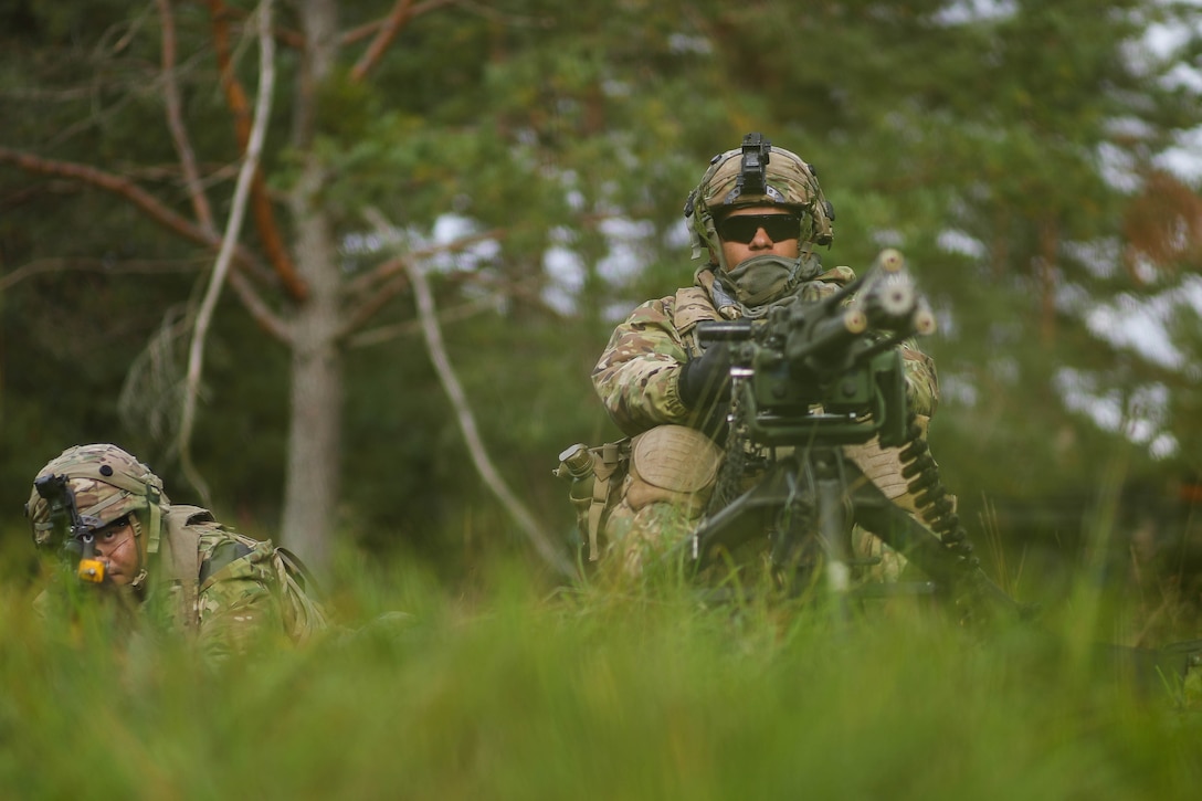 Two soldiers in camouflage uniforms crouch in tall grass and aim weapons.