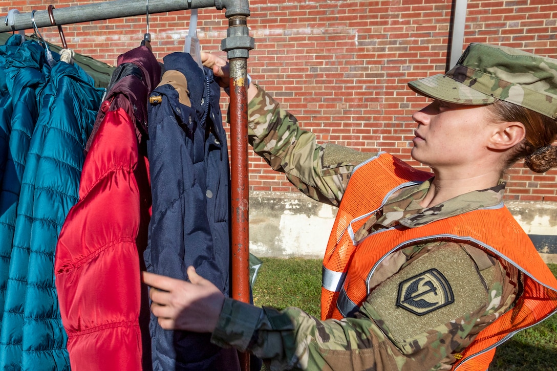 A soldier arranges winter coats on a clothing rack outside a brick building.