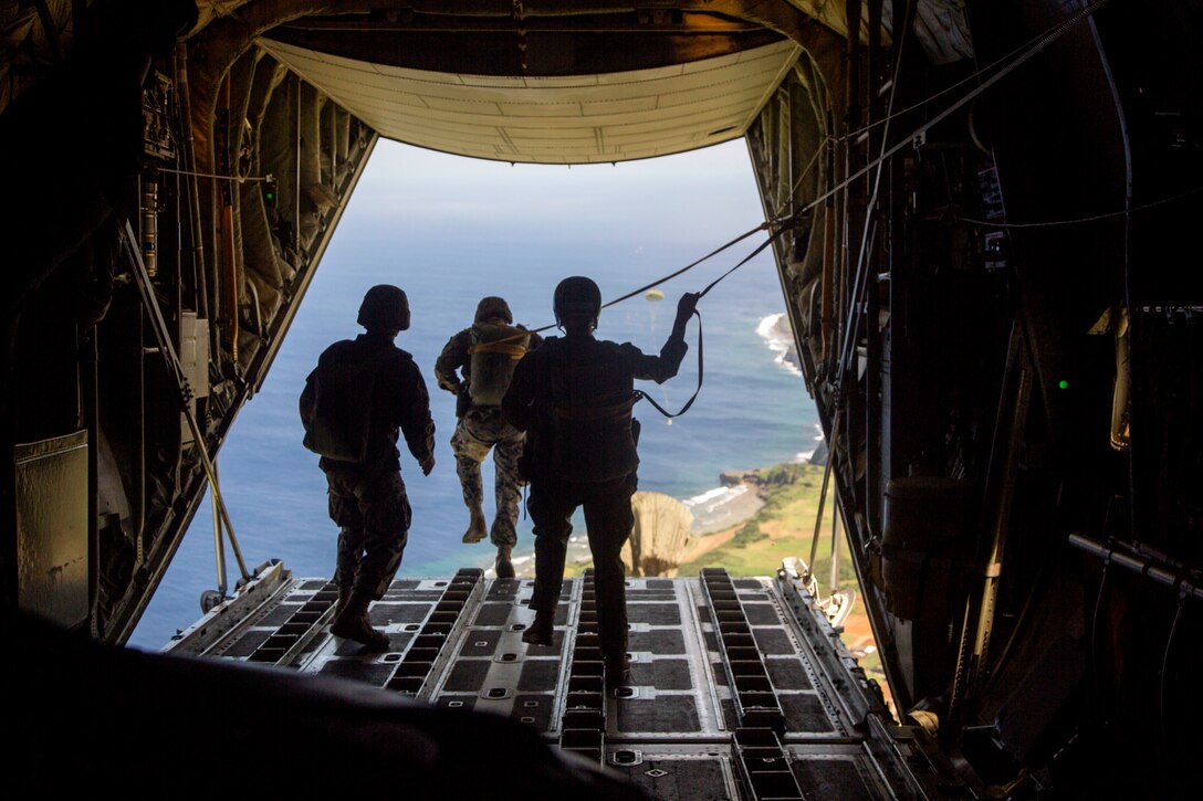 A Marine jumps out the back of an open aircraft over an island coastline as two others watch from inside the aircraft.