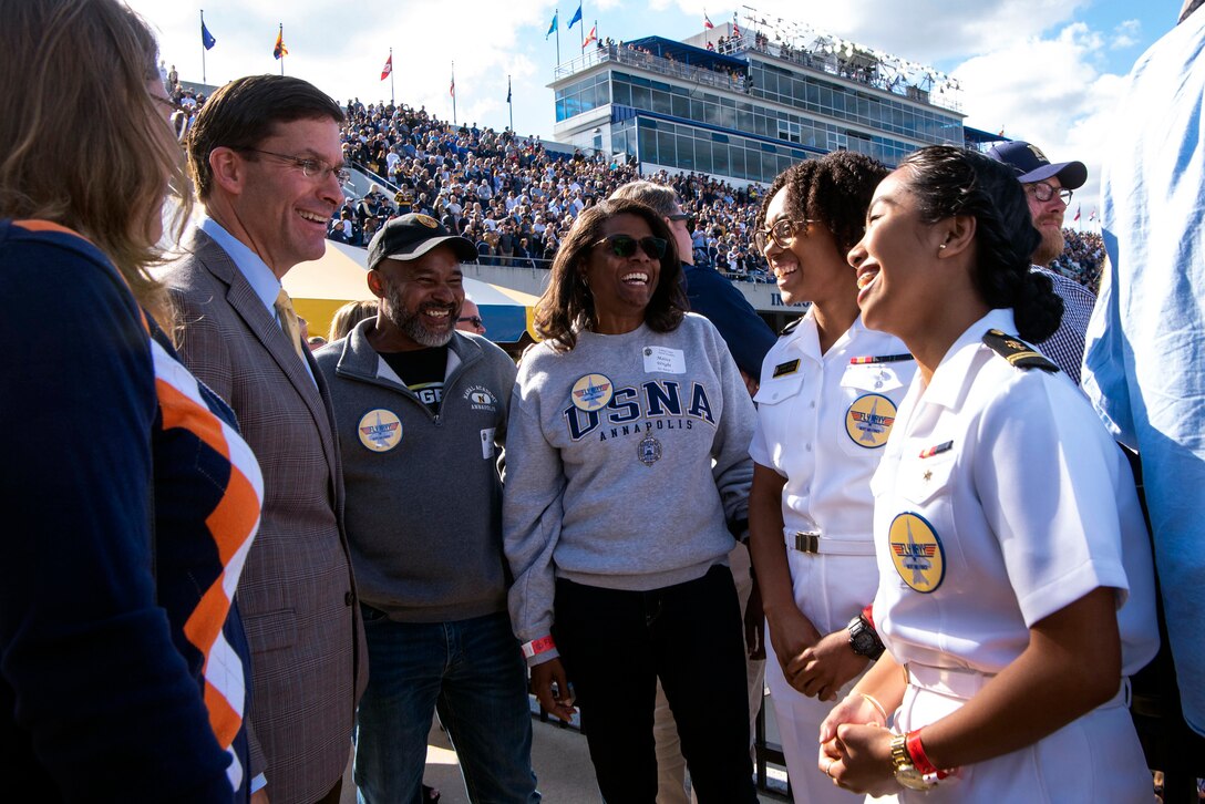 Women in navy uniforms speak with people standing in a circle, a crowded football stadium in the background.