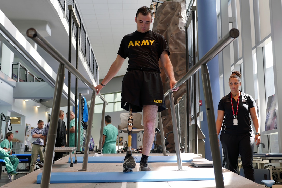 Man with a prosthetic leg who is wearing black shorts and a T-shirt reading “ARMY” walks along rails as physical therapist watches.