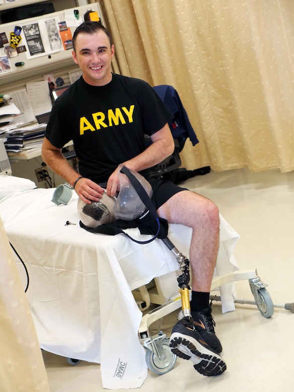 Man with a prosthetic leg wearing black shorts and a T-shirt reading “ARMY” sits on a rehabilitation facility bed.