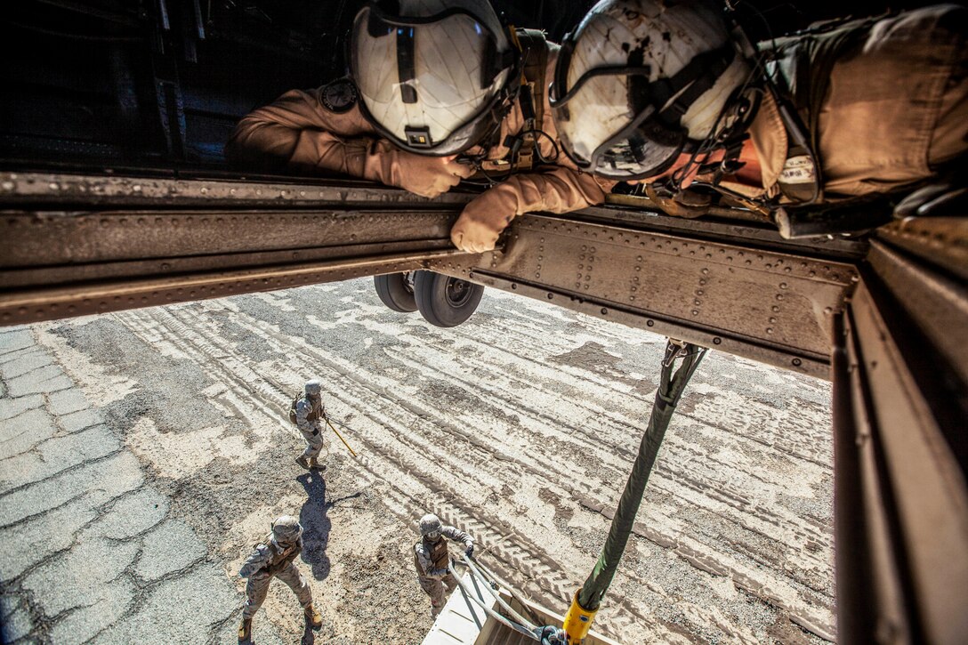 Marines in an aircraft look through a floor opening at Marines working on the ground.