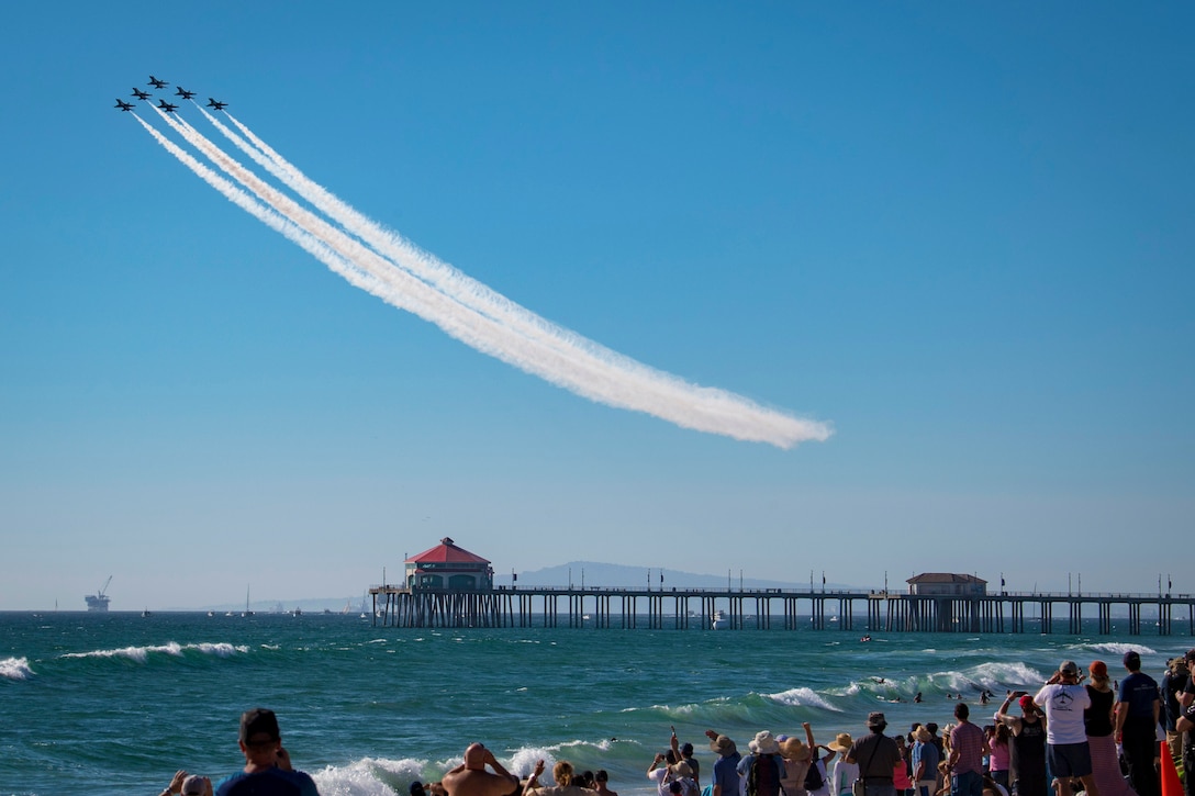 Six jets make trails together midair above a pier with spectators on the beach.