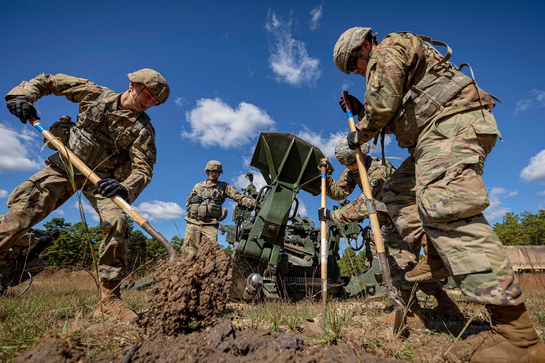 Soldiers dig a hole; some stand next to a howitzer.
