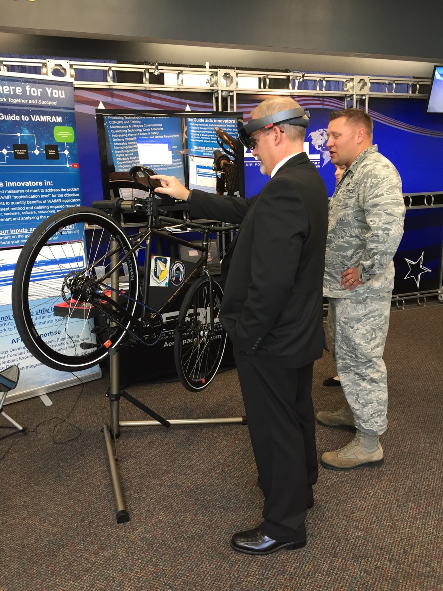 Dr. Michael Gregg (left), director of the Air Force Research Laboratory Aerospace Systems Directorate, uses a mixed-reality application to change the tire on a Wright Cycle Company bicycle as Capt. David Eisensmith guides the process. The demonstration, on display at the 2019 Air Vehicles Technology Symposium, is part of the Virtual, Augmented, and Mixed Reality for Aircraft Maintenance team’s effort to show the technology’s potential for future aircraft maintenance. (U.S. Air Force Photo)