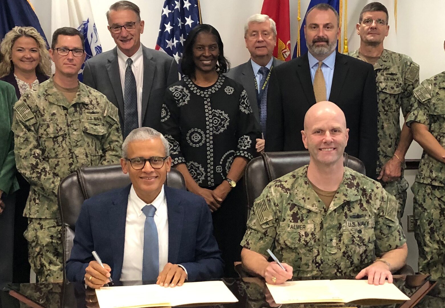 two men smile at camera while signing charter as group stands behind them.