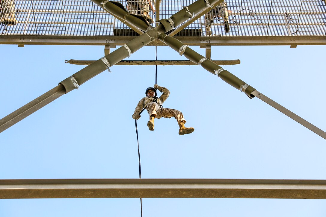 A Marine suspended from a rope hangs mid-air between triangular beams.