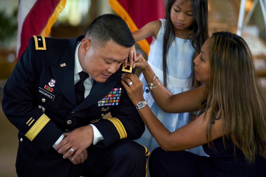 A soldier kneels as a woman and girl pin his rank to him.