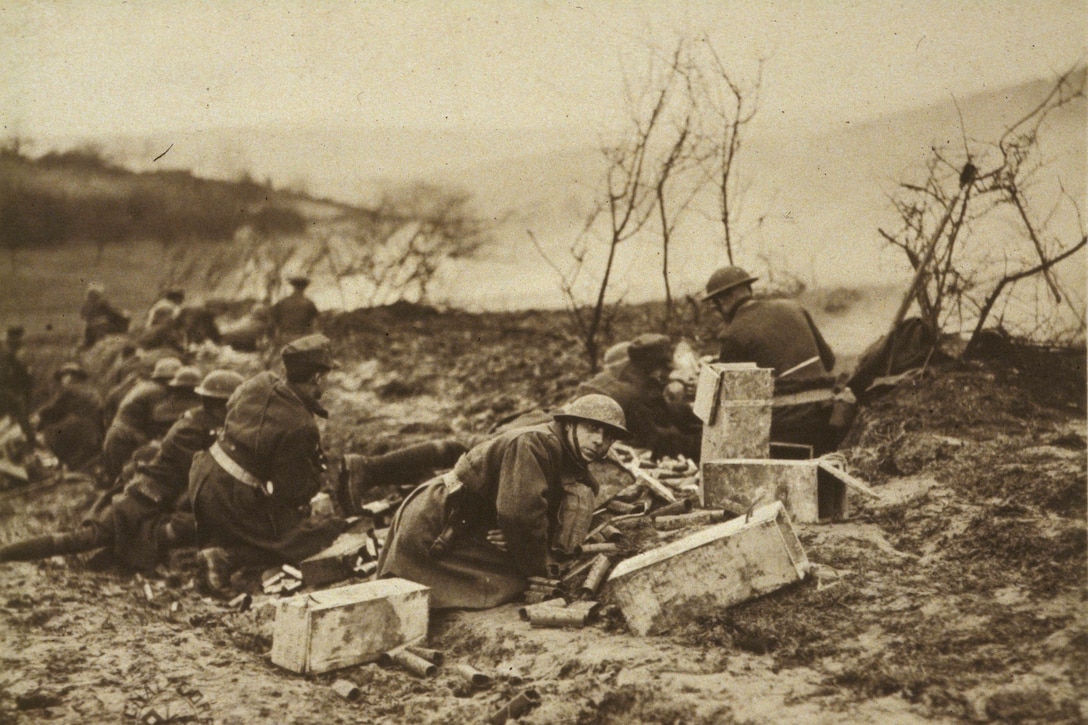 World War I soldiers ducking down low are surrounded by large cartridges as they battle in a barren wasteland. Smoke can be seen in the distance. One soldier looks directly at the camera.
