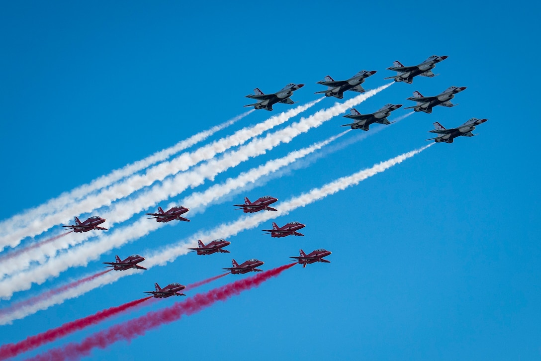 Two groups of military jets fly across a blue sky trailing red and white smoke.