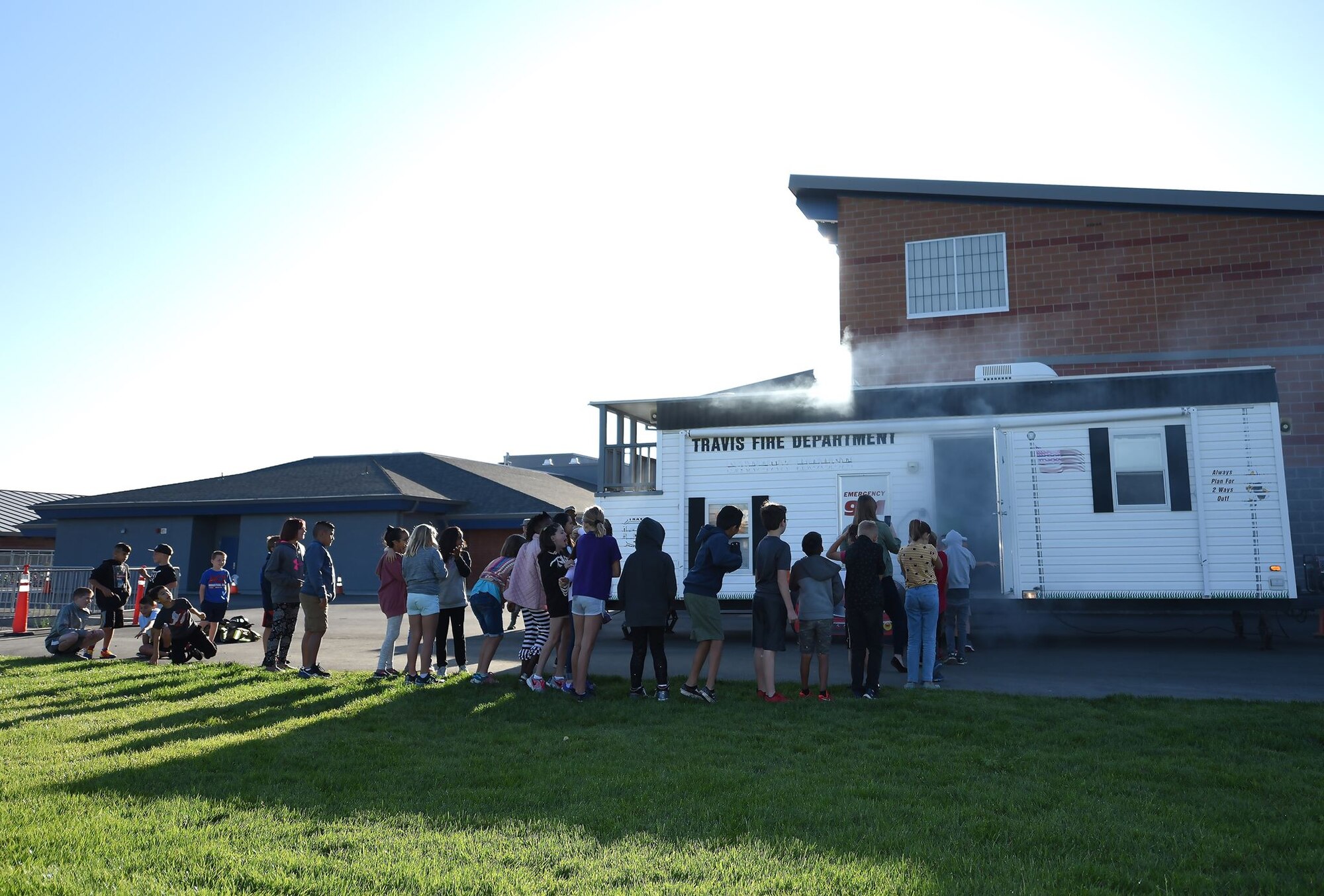 Grade-school aged children sit outside on a sunny morning next to a trailer filled with simulated smoke
