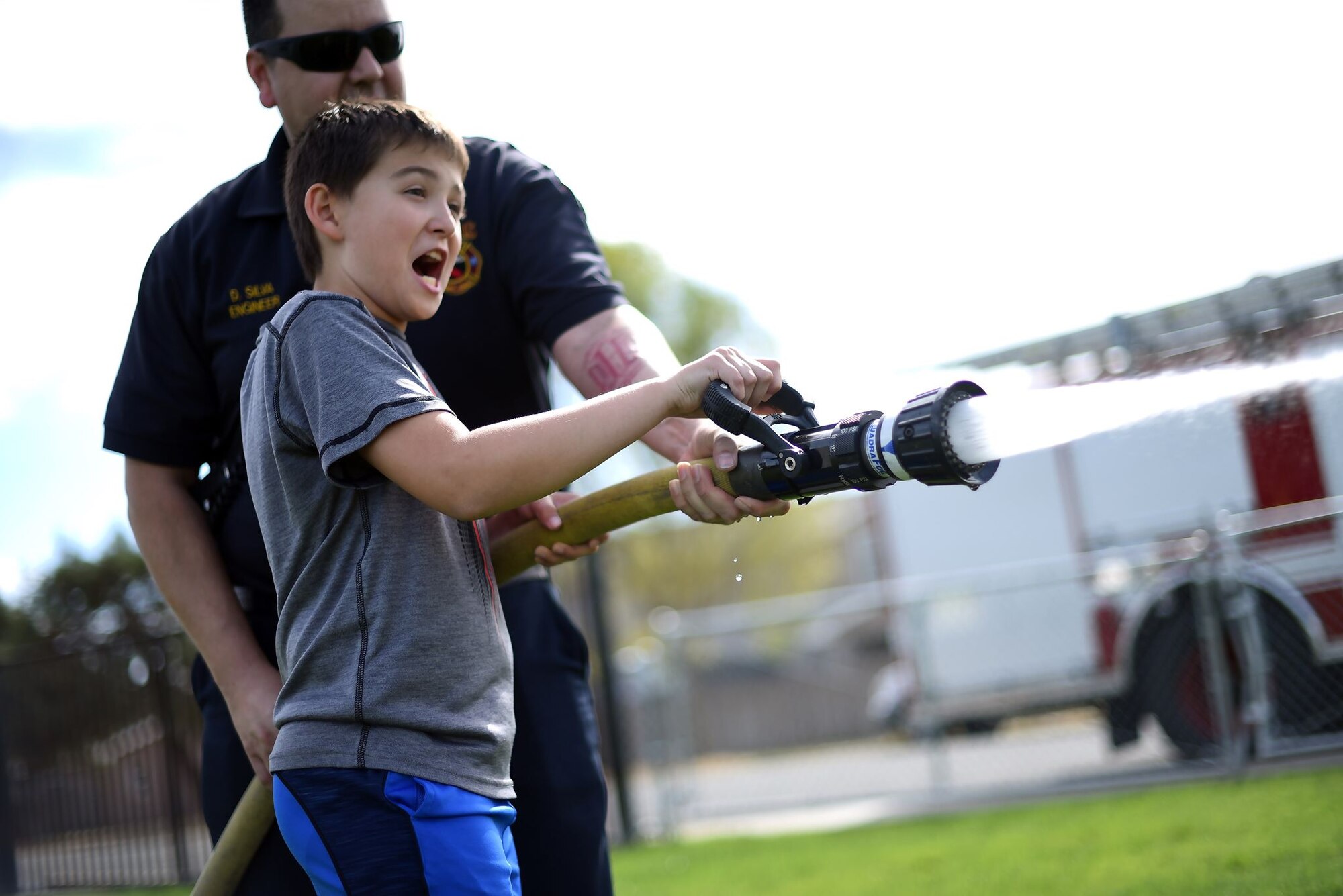 A grade school-aged boy is aided by a man in a firefighter uniform in using a fire hose to douse cardboard cutouts simulating fires.