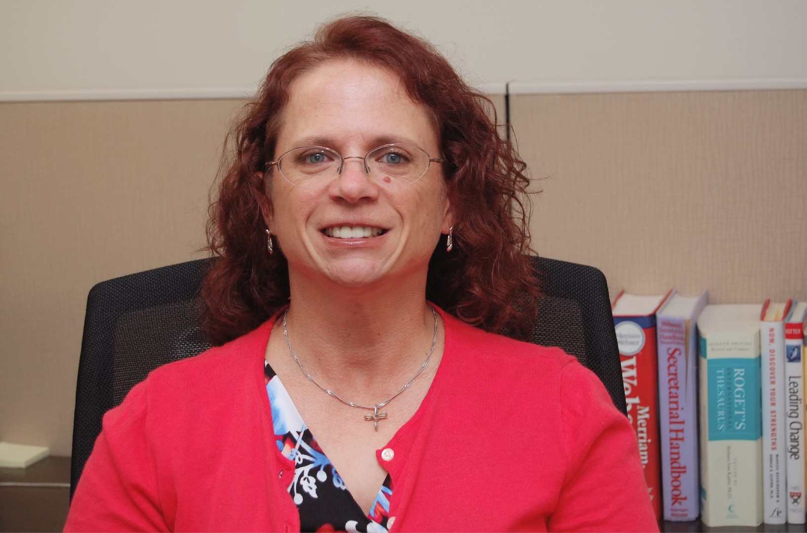 Smiling lady sits in a chair in her cubicle with books in the background.