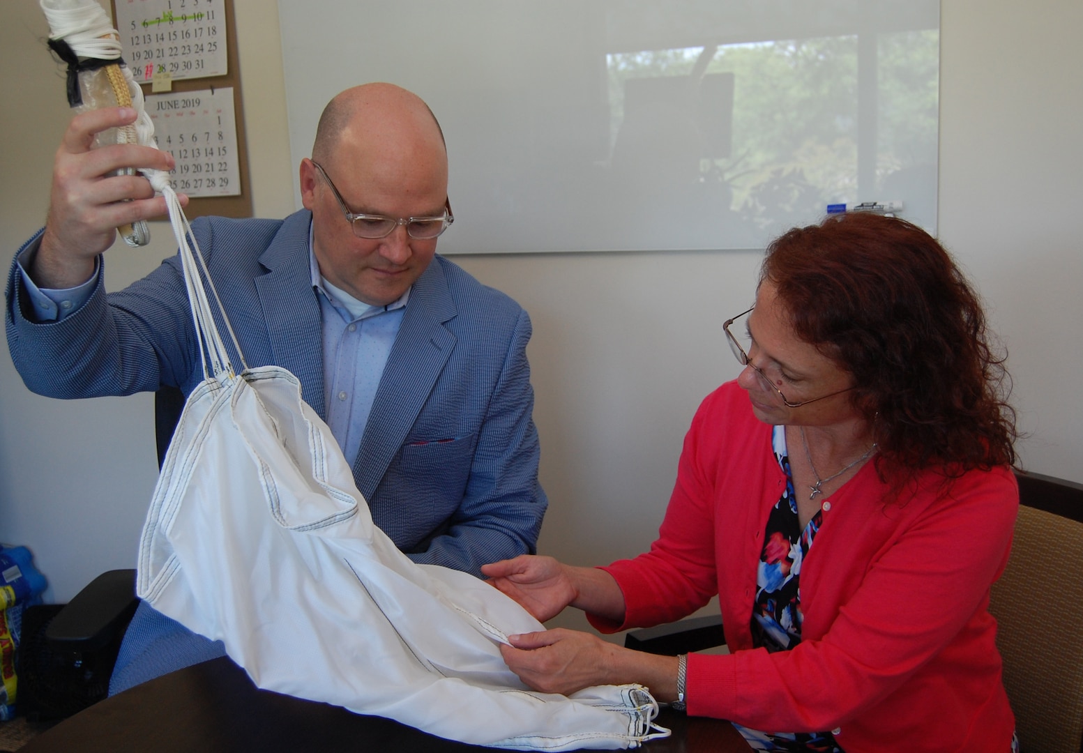 A male and female sit at a table in an office and inspect a parachute.