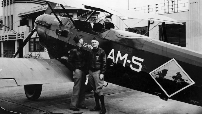 Two men stand in front of a prop aircraft.