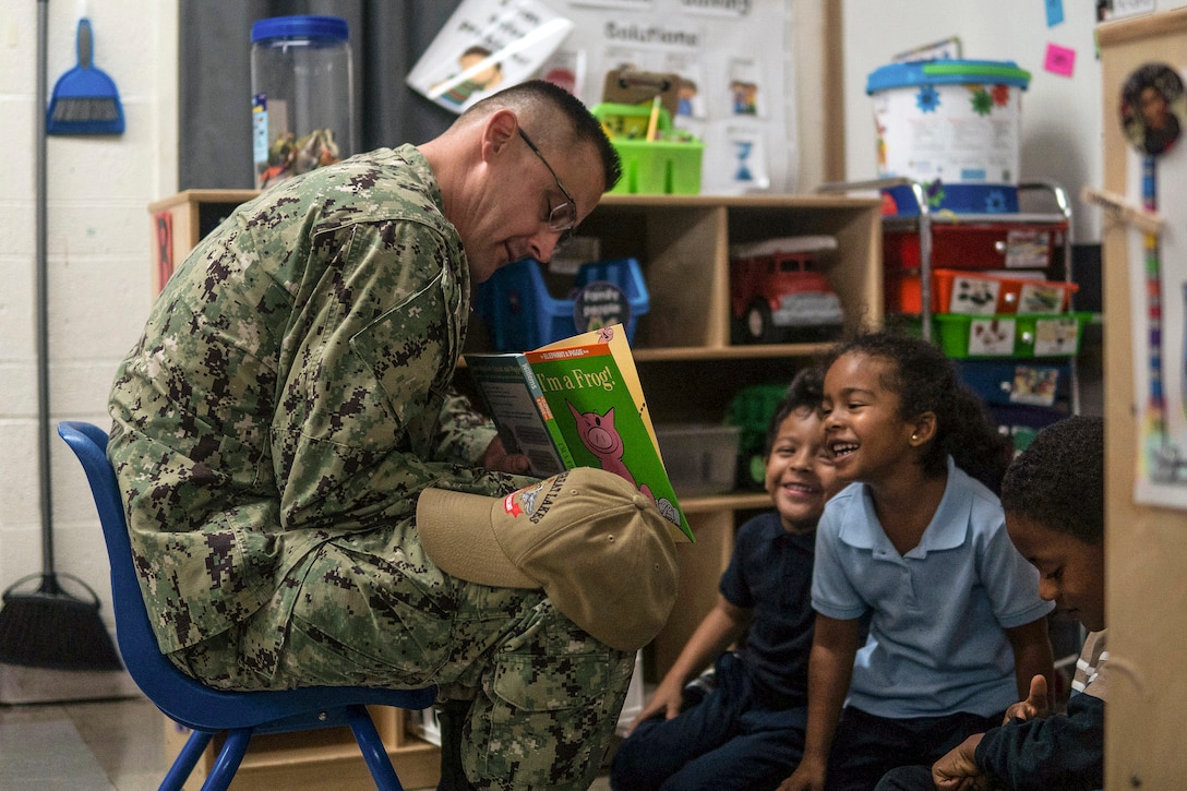 Children sitting on a classroom floor smile as a sailor reads to them.
