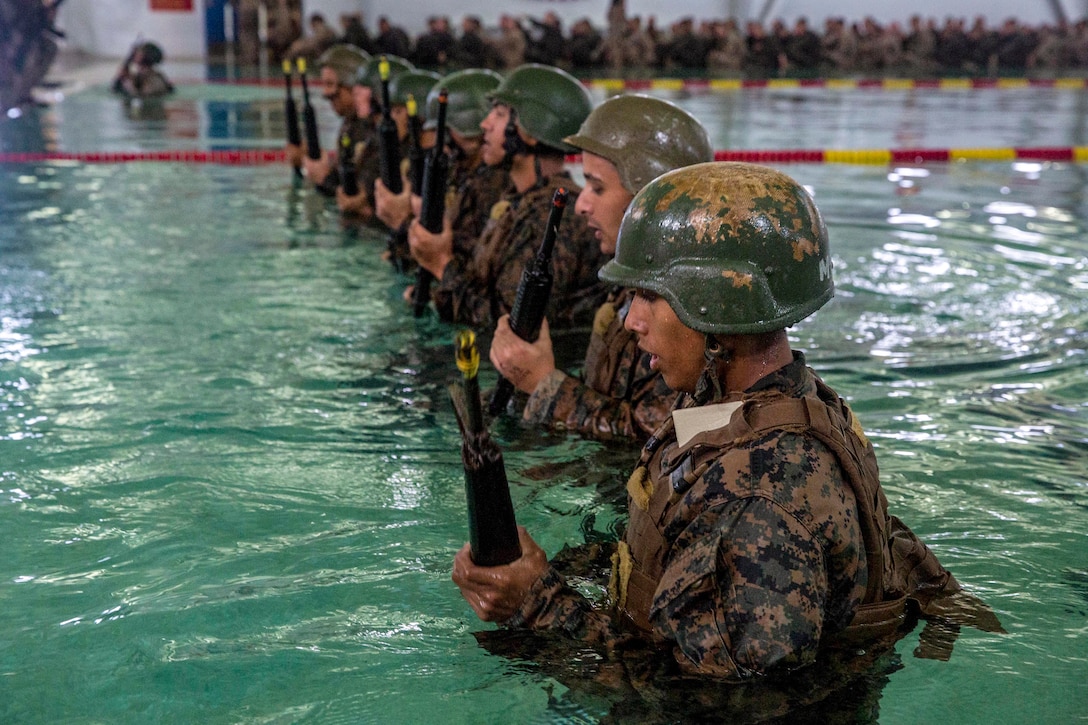 A group of Marine recruits stand in a pool, in a straight line, wearing their uniform and holding onto weapons.