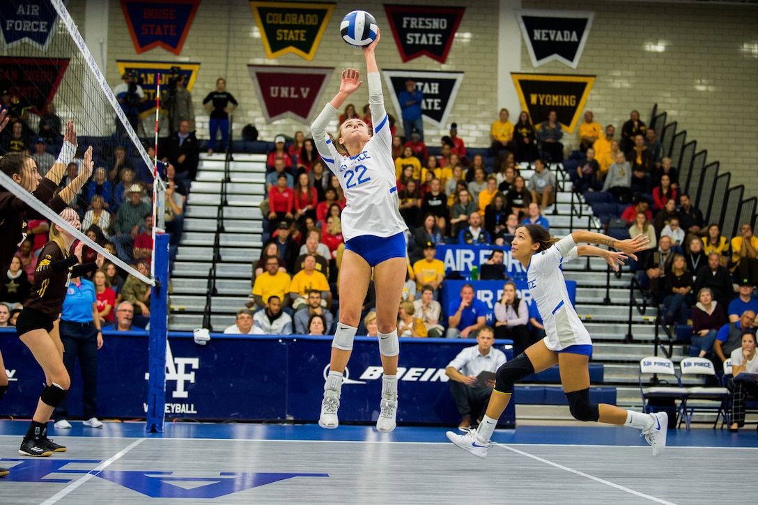 Two volleyball players jump in the air to spike a ball during a game while others look on.