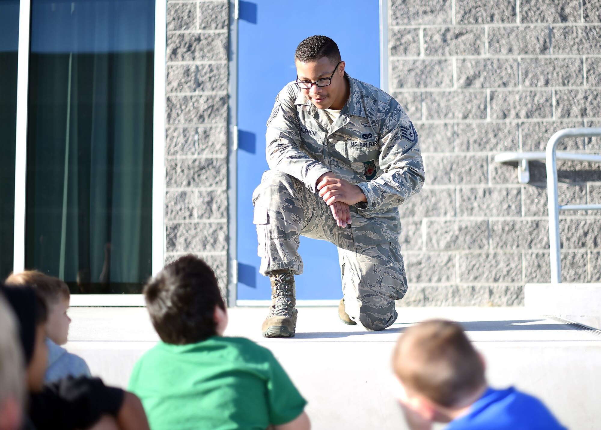 A uniformed member of the Air Force bends down on on knee to speak with grade-school aged children. It's sunny. The children listen to the Airman attentively.