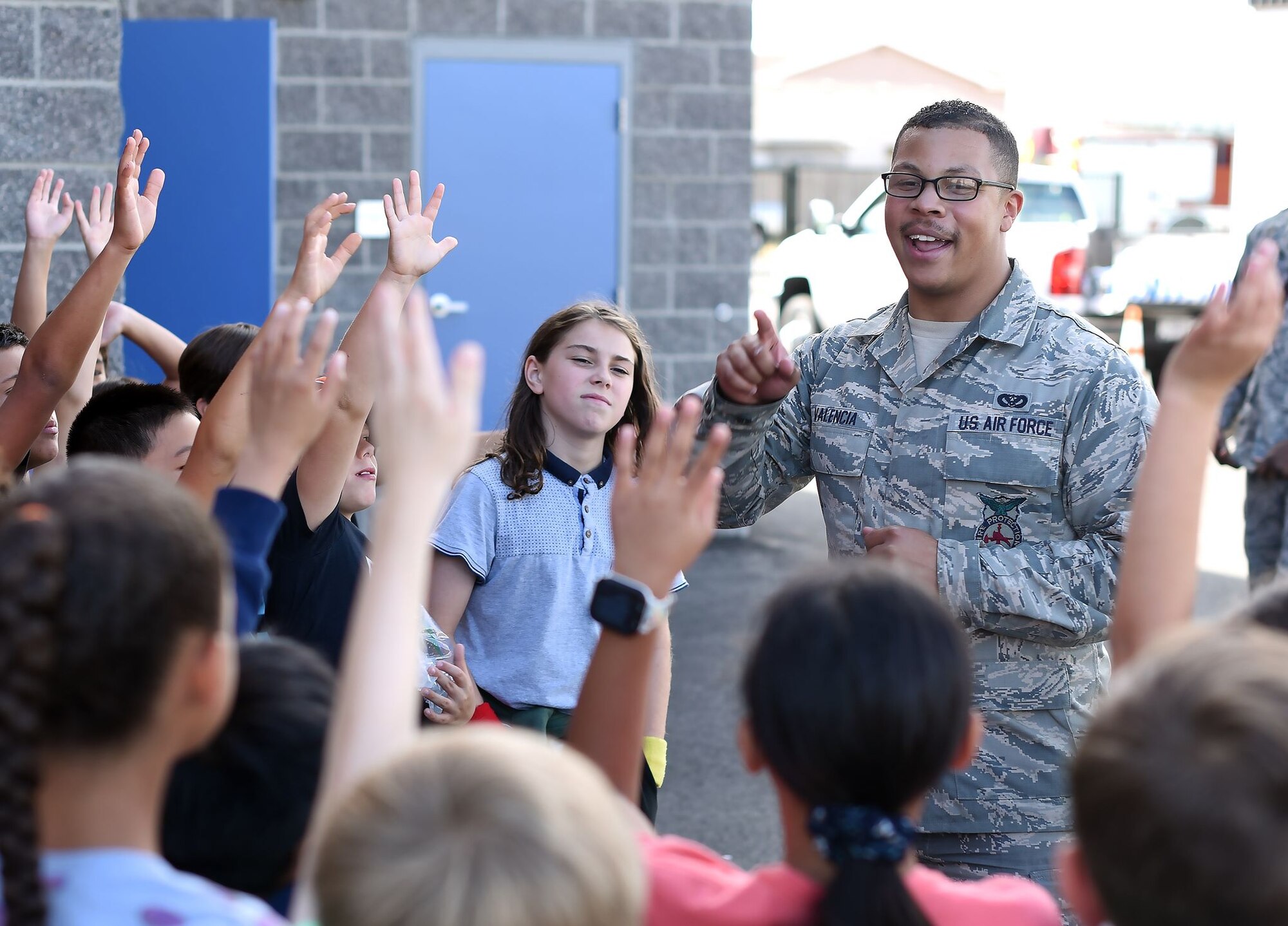 A uniformed member of the Air Force stands among grade school-aged children and look to be asking them a question. Many of the children's hands are up in gesture of a question or as a means of participation.